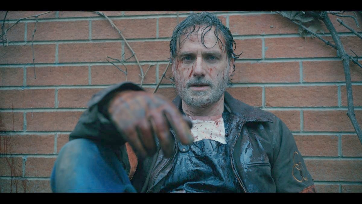 Rick Grimes sits against a wall with his right hand visible and blood on his face in the ones who live walking dead spinoff